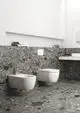 Moderne Badezimmeransicht mit grauem Mosaikboden und -wand. Ideal Standard Toiletten sowie Bidet in Weiß, schlichte Ablage mit Hygieneartikeln im Hintergrund.